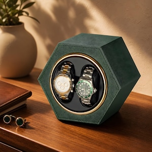 May include: A hexagonal watch winder box covered in dark green textured material with a gold-toned circular frame. The interior holds two wristwatches, one with a white face and gold-silver band, and one with a green face and silver band.