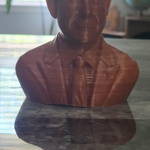 May include: A brown bust of a man wearing a suit and tie. The sculpture is 3D-printed and has a textured surface. The bust is sitting on a reflective surface.