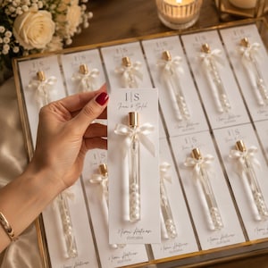 May include: A collection of clear glass tubes filled with white pearls, each attached to a white card featuring the text I|S Irem & Salih and Nisan Hatirasi 25/07/2026. Each tube is topped with a gold cap and a small white satin ribbon bow.
