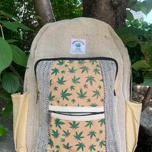 May include: Beige hemp backpack with a front pocket featuring a pattern of green cannabis leaves on a tan background. The backpack has side pockets and a zipper closure. The top of the backpack has a label that says "Himalayan Hemp".