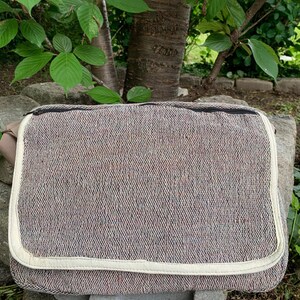 May include: A rectangular messenger bag with a woven brown and cream fabric. The bag has a cream-colored border and a black zipper. A tan strap is attached to the side of the bag. The bag is sitting on a stone surface.