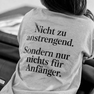 以下が含まれることがあります： 背面に「Nicht zu anstrengend. Sondern nur nichts für Anfänger.」という黒い文字がプリントされた明るい色のTシャツ。クルーネックと半袖のデザインで、背後から見た様子が写されています。