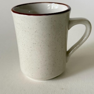 Puede incluir: Una taza cilíndrica de cerámica con un acabado moteado en color blanco roto y un borde marrón oscuro. La taza presenta un asa curva y lisa en el lado derecho y un diseño sencillo y limpio. El interior es blanco y el exterior tiene textura.
