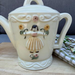 May include: A cream-colored ceramic teapot featuring a raised relief design of a figure in a skirt holding two yellow flowers. The teapot has a matching lid with a handle, a curved spout, and a decorative embossed border around the top and base.