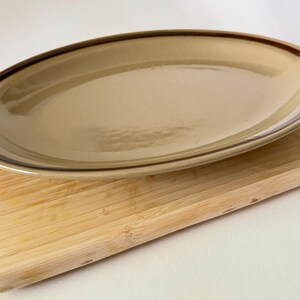 May include: An oval-shaped ceramic serving platter with a smooth, glossy tan finish and a dark brown band encircling the rim. The platter features a shallow basin and a slightly raised edge, presenting a clean, functional design for food service.