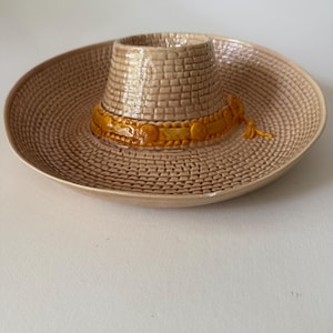 Könnte beinhalten: Eine beige, sombrero-förmige Keramikschale mit einem strukturierten Flechtmuster auf der Krempe und der Krone. Ein glänzendes, erhabenes orangefarbenes Band mit kreisförmigen Akzenten umschließt die Basis der Krone, ergänzt durch eine kleine Quaste.