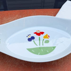 May include: A white oval ceramic serving dish with integrated handles on both ends. The interior features a central graphic of a green semi-circle base with stylized red, yellow, and blue flowers, and smaller matching floral motifs on each side.