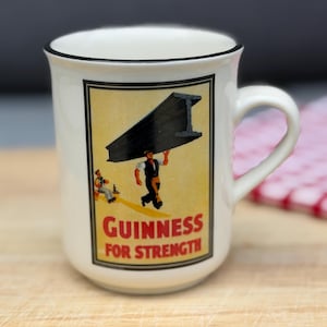 May include: A white ceramic mug with a black rim and a handle, featuring a vintage-style graphic of a person carrying a large steel beam on their shoulder. The text "GUINNESS FOR STRENGTH" is printed in red and black letters below the illustration.