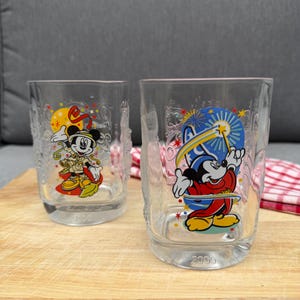 Può includere: Due bicchieri in vetro trasparente con grafiche di Mickey Mouse. Un bicchiere mostra Mickey in tenuta da safari con un sole giallo, l'altro ritrae Mickey come uno stregone con cappello blu e bacchetta. Entrambi hanno l'anno 2000 in rilievo.