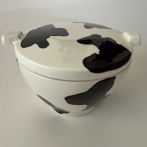 May include: A white ceramic bowl with a lid, featuring an irregular black cow-print pattern. The bowl has two small, protruding handles on opposite sides, and the lid sits flush with the rim, mirroring the black and white spotted design.
