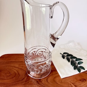 Puede incluir: Una jarra de cristal transparente con un cuerpo alto y esbelto, un asa curva y un pico vertedor ancho. La base de la jarra está decorada con un patrón circular en relieve que se enrolla alrededor de la sección inferior del recipiente.