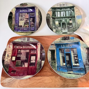 May include: Four round ceramic plates featuring detailed illustrations of French storefronts. The designs include a purple shop labeled EMILIE, a green restaurant labeled Chez JEROME, a red wine shop labeled VINS DE BOURGOGNE, and a blue bookstore labeled LIVRES.