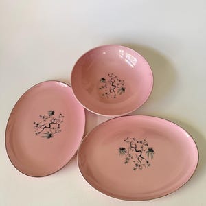Op de afbeelding: Een set van drie lichtroze keramische schalen bestaande uit één ronde kom en twee ovale schalen. Elk stuk is voorzien van een centrale zwart-witte grafische illustratie van een dennentak met naalden en een dunne zilveren metalen rand.