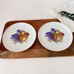 Op de afbeelding: Twee ronde witte porseleinen borden met een gouden rand, elk voorzien van een centrale illustratie van een bruine peer, een tros paarse druiven en een enkele rode aardbei met groene bladeren, geplaatst op een bruine houten plank.