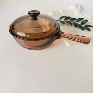 Pode incluir: Uma panela de vidro translúcido na cor âmbar com tampa a condizer e uma pega única. O utensílio de cozinha apresenta uma forma de taça lisa e arredondada e uma tampa circular com um pequeno puxador, fabricado em vidro castanho.