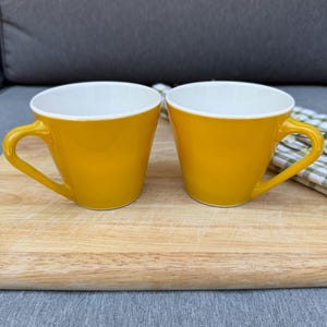 Puede incluir: Dos tazas a juego con un exterior amarillo vibrante y un interior blanco liso. Cada taza presenta un borde ancho y acampanado, un cuerpo cónico y un asa curva en el lateral. Las tazas tienen un acabado brillante y un diseño funcional.
