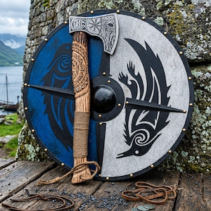 Può includere: Uno scudo rotondo in legno con un design diviso in blu e grigio, motivi di corvi neri e un umbone centrale in metallo, abbinato a un'ascia in legno intagliato con testa in metallo incisa e manico avvolto.