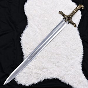 Peut inclure: Une longue épée en acier à double tranchant dotée d'une poignée en bronze métallique avec des détails complexes en forme de tête de dragon sur le pommeau et la garde, ornée d'une petite pierre rouge centrale.