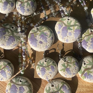 Puede incluir: Una colección de colgantes y pendientes de piedra redondos y moteados con flores de glicinia moradas pintadas a mano sobre ramas verdes. Las piezas de joyería están ensartadas en cordones de cuentas transparentes, blancas y moradas.