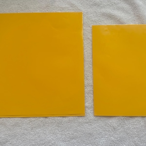 Op de afbeelding: Twee rechthoekige, effen gele vellen papier liggen op een getextureerd, lichtgrijs oppervlak. Het grotere vel is vierkant, terwijl het kleinere rechthoekig is. De vellen hebben een levendige, effen gele kleur.