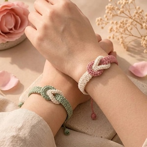 May include: Two woven cord bracelets featuring a central knot design. One bracelet combines dusty rose and cream threads, while the other features sage green and cream threads. Each bracelet has a simple adjustable cord closure with a small bead.