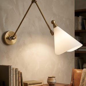 May include: A brass wall-mounted lamp with an adjustable arm and a white, triangular shade. The lamp is mounted on a light-colored wall. The lamp is next to a wooden shelf with books and a small ceramic pot.
