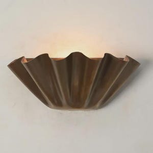 May include: A bronze-colored wall sconce with a ruffled, seashell-like design. The light fixture is mounted on a white wall and emits a warm glow. The sconce's unique shape adds a decorative element to the space.