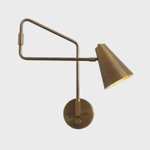 May include: A brass-toned wall sconce with a conical shade and adjustable arm. The sconce features a circular base and a geometric arm design, ideal for modern or industrial decor. The light is on.
