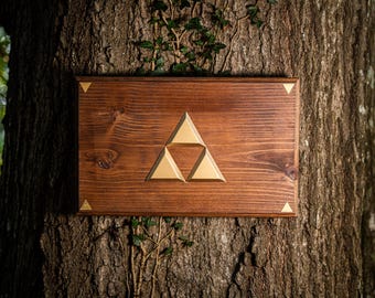 May include: A rectangular wooden plaque featuring a central gold-colored Triforce emblem composed of three triangles. The plaque includes four smaller gold-colored triangles at each corner, set against a dark brown wood grain finish.