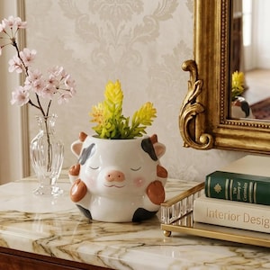 May include: A white ceramic cow-shaped planter with black and brown spots, holding yellow and green plants. The planter has a smiling face and is on a marble surface. A small glass vase with pink flowers is to the left.