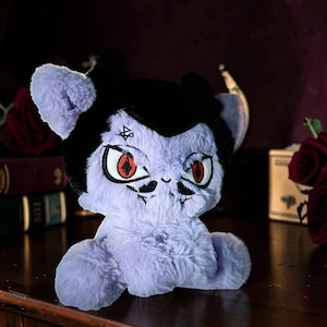 May include: A plush toy featuring a light purple, fuzzy body with black hair and pointed ears. The face has embroidered red eyes with black pupils, a small mouth, and a black symbol on the forehead. The character has a vampire-like appearance.