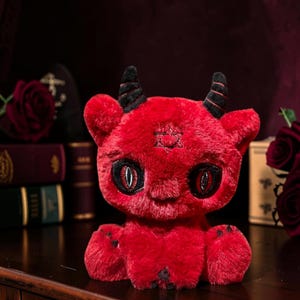 May include: A plush toy shaped like a small red devil with soft fur, two black horns, and large eyes with vertical pupils. A six-pointed star symbol is embroidered on its forehead. The creature has small paws with black claws and a rounded body.