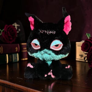 May include: A plush toy featuring a black cat-like creature with large pointed ears, bright pink inner ear accents, and teal fur on the lower face. The creature has embroidered eyes with light blue lids and pink irises, and small pink paw pads.