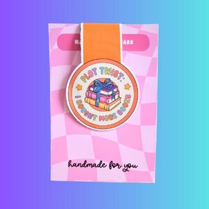 Peut inclure: Un marque-page coloré avec un design rond représentant une pile de livres liés par un ruban bleu. Le texte dit "PLOT TWIST: I BOUGHT MORE BOOKS". Le marque-page est fixé à une carte rose et orange avec le texte "handmade for you".