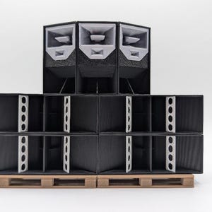 Op de afbeelding: Een zwart en grijs luidsprekersysteem met gestapelde rechthoekige dozen en schuine luidsprekers aan de bovenkant. Het systeem staat op een houten pallet tegen een witte achtergrond. De luidsprekers hebben witte accenten.