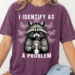 May include: A dark purple short-sleeve t-shirt featuring a black and white graphic of a raccoon holding a drink cup. The text "I IDENTIFY AS A PROBLEM" is printed in white above and below the raccoon, surrounded by several small butterflies.