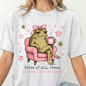 May include: A light grey t-shirt featuring a graphic of a green toad wearing a pink bow, sitting in a pink armchair while holding a pink mug. The text reads, first of all, Jesus thinks i'm a delight, surrounded by small pink and gold stars.