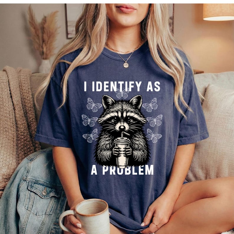May include: A navy blue short-sleeve t-shirt featuring a graphic of a raccoon holding a cup with a straw. The text "I IDENTIFY AS A PROBLEM" is printed in white above and below the raccoon, surrounded by several white butterfly illustrations.