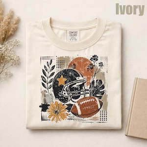 May include: An ivory crew neck t-shirt with a Comfort Colors label. The graphic features a black football helmet with a gold star, a brown football, a yellow flower, and botanical elements against a rust-colored circle and abstract shapes.