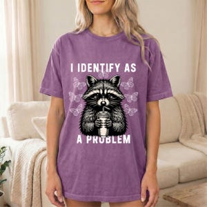 May include: A purple short-sleeve t-shirt with a graphic print of a raccoon holding a cup with a straw. The text I IDENTIFY AS A PROBLEM is printed in white above and below the raccoon, surrounded by several small white butterfly outlines.