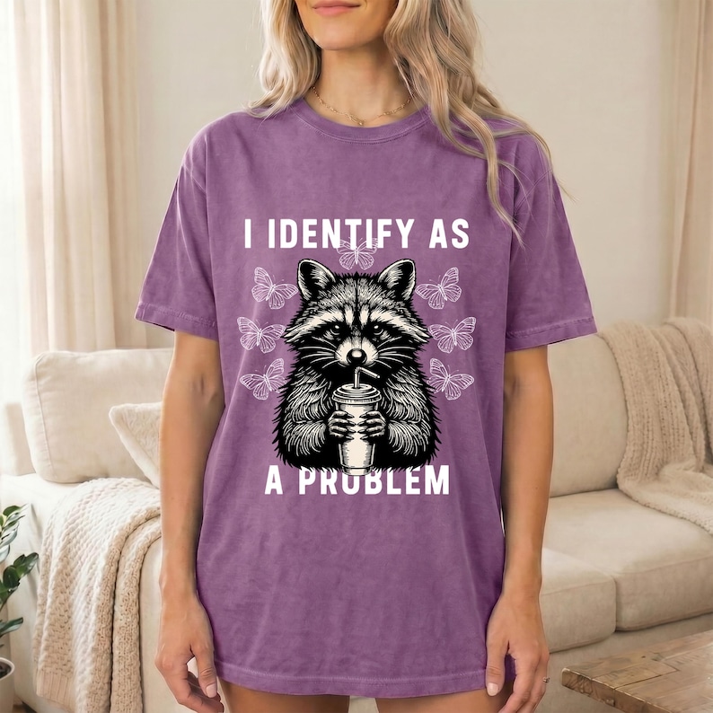 May include: A purple short-sleeve t-shirt with a graphic print of a raccoon holding a cup with a straw. The text I IDENTIFY AS A PROBLEM is printed in white above and below the raccoon, surrounded by several small white butterfly outlines.