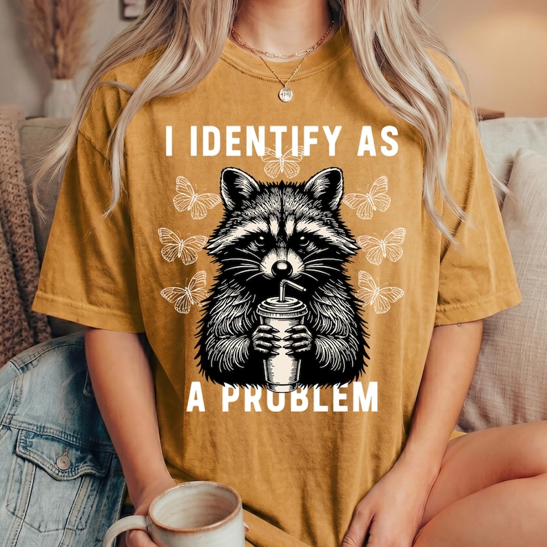May include: A mustard yellow short-sleeve t-shirt featuring a black and white graphic of a raccoon holding a drink cup. The text "I IDENTIFY AS A PROBLEM" is printed in white above and below the raccoon, surrounded by white butterfly illustrations.