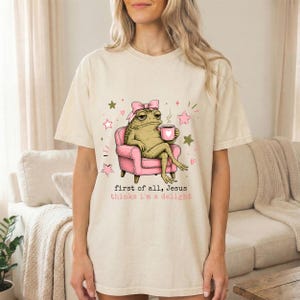 May include: A cream-coloured t-shirt featuring a graphic of a green toad wearing a pink bow, sitting in a pink armchair while holding a pink mug with a white heart. The text reads, first of all, Jesus thinks i'm a delight, surrounded by small stars.