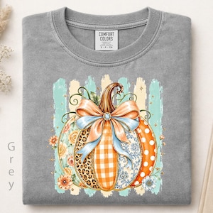 May include: A grey crewneck t-shirt featuring a graphic of a pumpkin decorated with various patterns including leopard print, orange gingham, and blue floral designs. A large light blue bow sits atop the pumpkin, which is set against light blue brushstrokes.