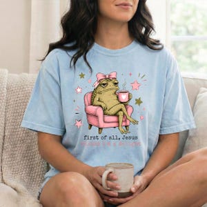 May include: A light blue t-shirt featuring a graphic of a green toad wearing a pink bow, sitting in a pink armchair while holding a pink mug. The text reads, "first of all, jesus thinks i'm a delight" in black and pink lettering with small stars.