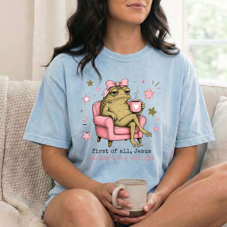 May include: A light blue t-shirt featuring a graphic of a green toad wearing a pink bow, sitting in a pink armchair while holding a pink mug. The text reads, "first of all, jesus thinks i'm a delight" in black and pink lettering with small stars.
