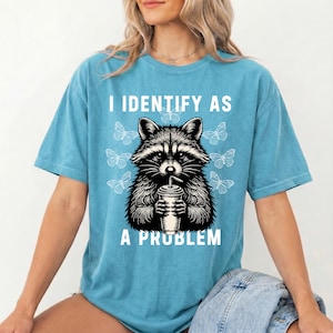 May include: A blue short-sleeve t-shirt featuring a black and white graphic of a raccoon holding a drink cup. The text "I IDENTIFY AS A PROBLEM" is printed in white above and below the raccoon, surrounded by several white butterfly illustrations.