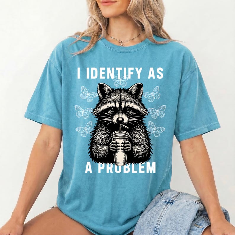 May include: A blue short-sleeve t-shirt featuring a black and white graphic of a raccoon holding a drink cup. The text "I IDENTIFY AS A PROBLEM" is printed in white above and below the raccoon, surrounded by several white butterfly illustrations.