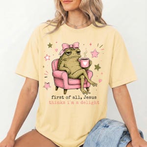 May include: A pale yellow t-shirt featuring a graphic of a green toad wearing a pink bow, sitting in a pink armchair while holding a pink mug. The text reads, "first of all, Jesus thinks i'm a delight," surrounded by small pink and green stars.