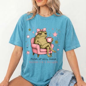 May include: A blue short-sleeve t-shirt featuring a graphic of a toad wearing a pink bow, sitting in a pink armchair while holding a pink mug with a heart. The text reads, first of all, Jesus thinks i'm a delight, surrounded by small pink and gold stars.
